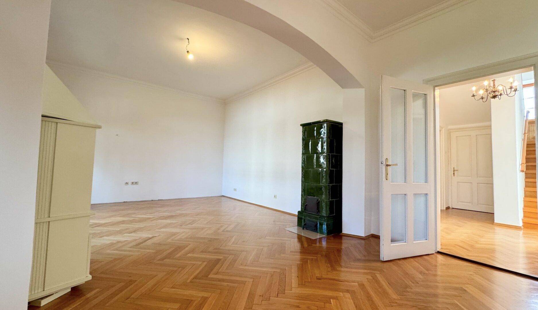 Großes Wohnzimmer mit Fischgrätparkett, einem grünen Kachelofen und Zugang zu weiteren Räumen.