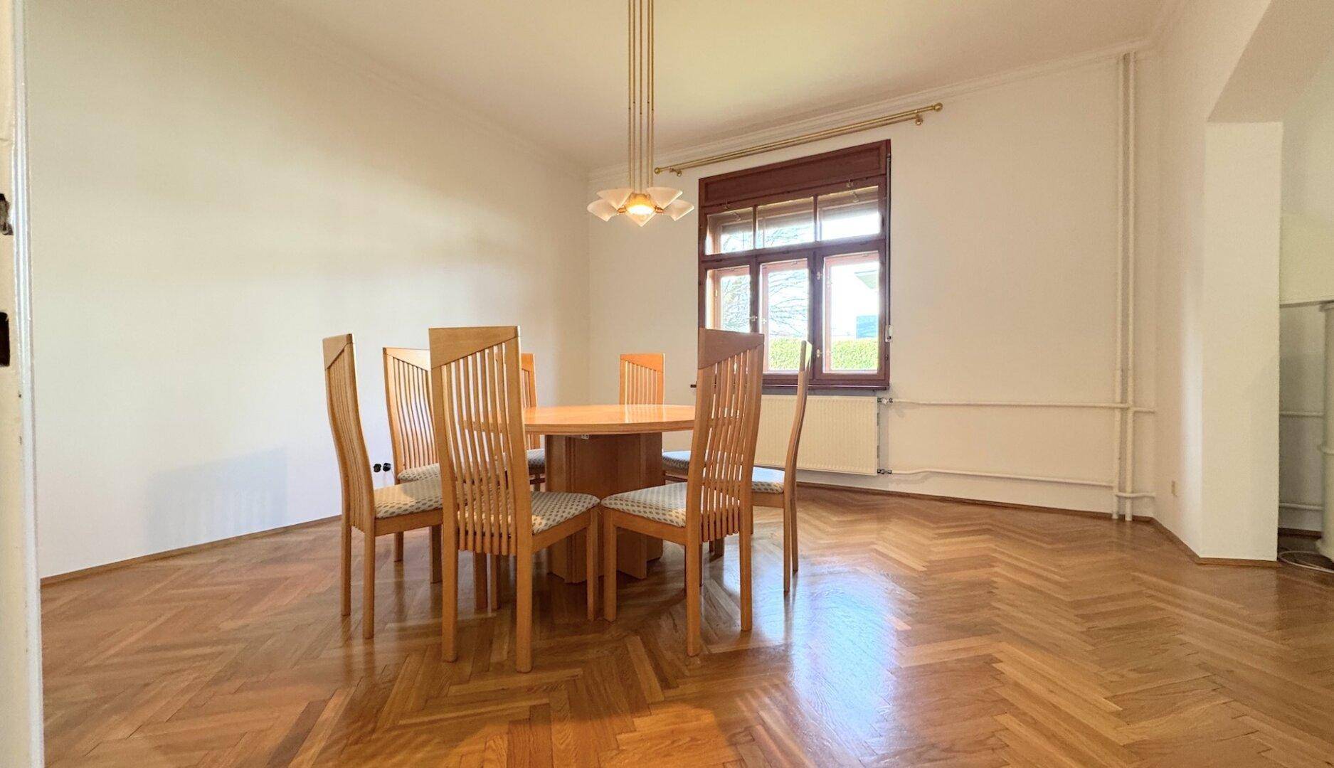 Heller Essbereich mit Fischgrätparkett, einem Holztisch und passenden Stühlen unter einem Fenster.