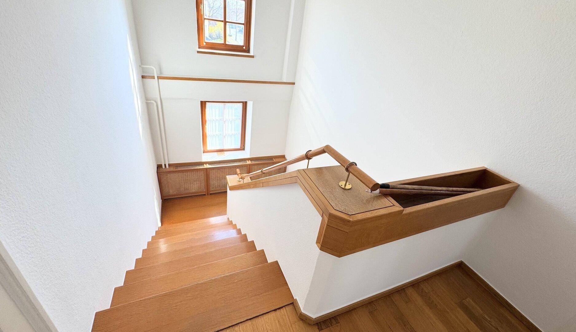Holztreppe mit Geländer und Fenstern, die den oberen Flur mit Tageslicht versorgen.