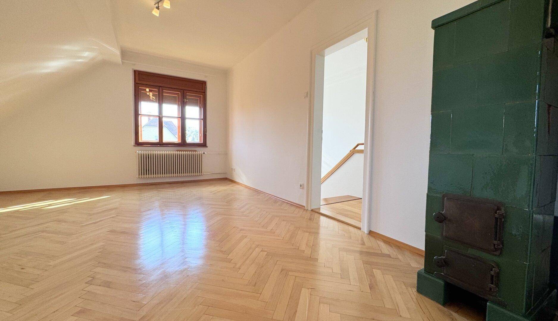 Geräumiges Zimmer im Obergeschoss mit Fischgrätparkett, Kachelofen und einem Fenster mit Blick nach draußen.