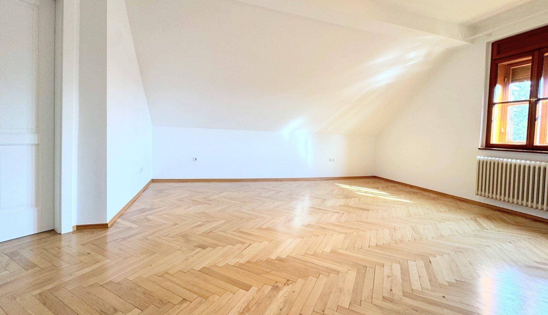 Großes, helles Zimmer mit Fischgrätparkett, Dachschrägen und einem Fenster, das viel Licht hereinlässt.