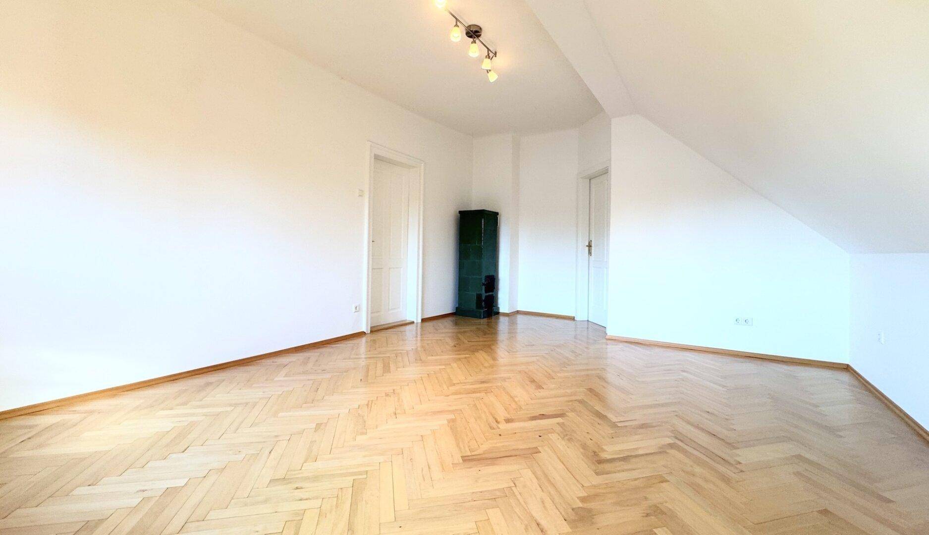 Helles Zimmer mit Fischgrätparkettboden, weißen Wänden und einem Kachelofen in der Ecke.