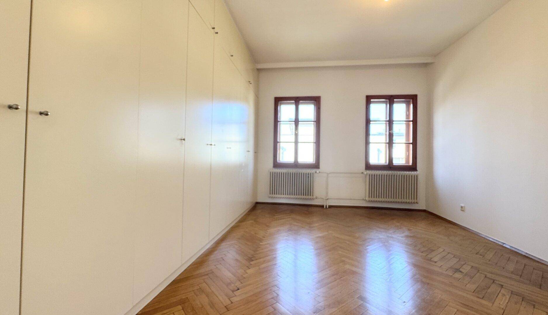Schlafzimmer mit Fischgrätparkett, Einbauschränken entlang einer Wand und zwei Fenstern.