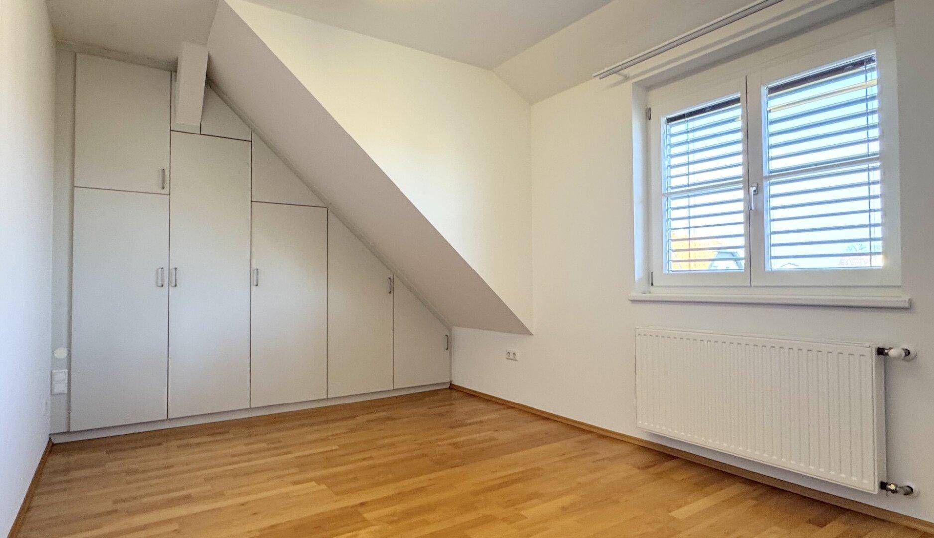 Helles Zimmer mit Holzparkett, Dachschräge und maßgefertigten Einbauschränken unter dem Fenster.