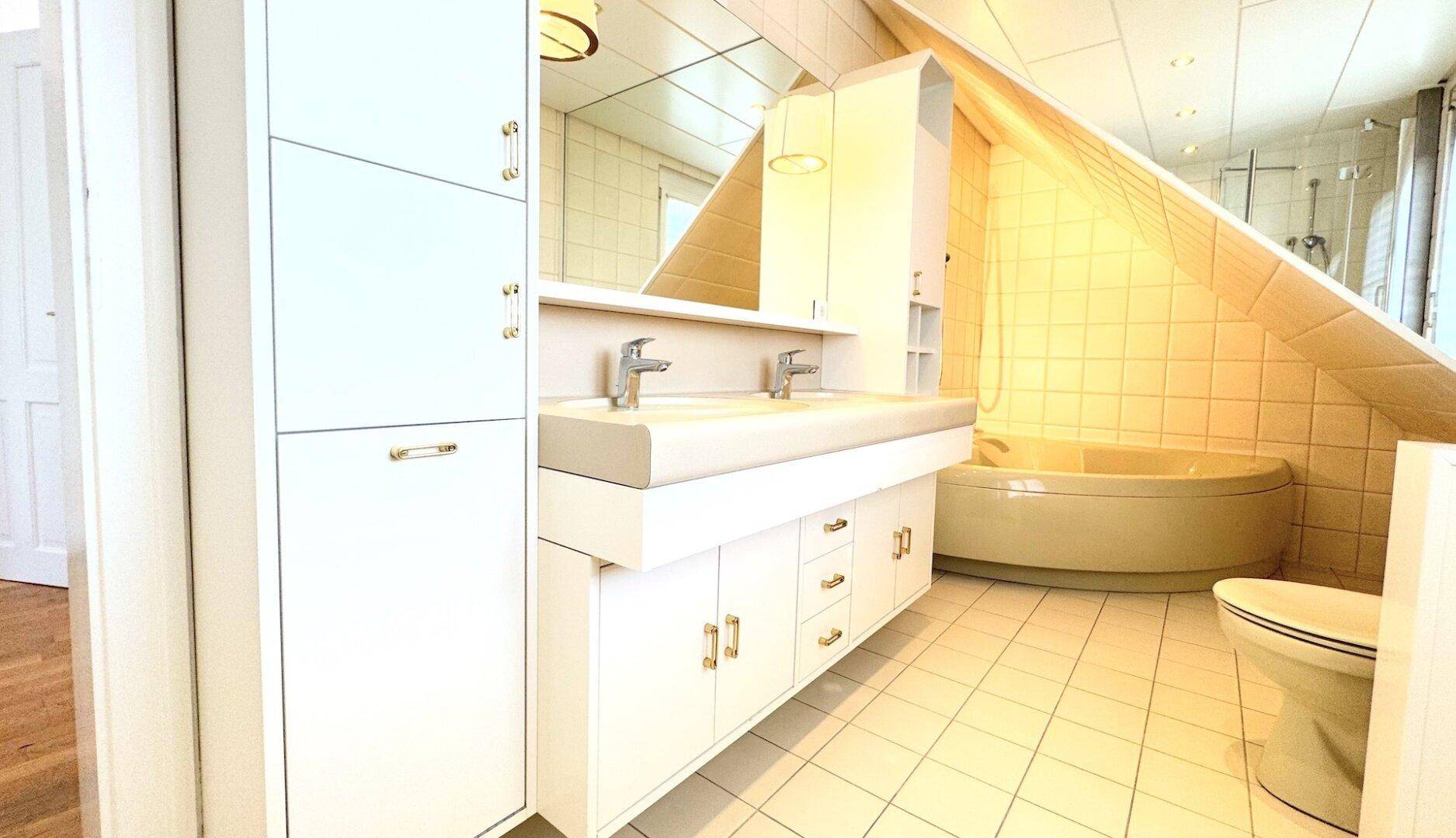 Badezimmer mit Doppelwaschbecken, Badewanne unter der Dachschräge und hellen Fliesen.