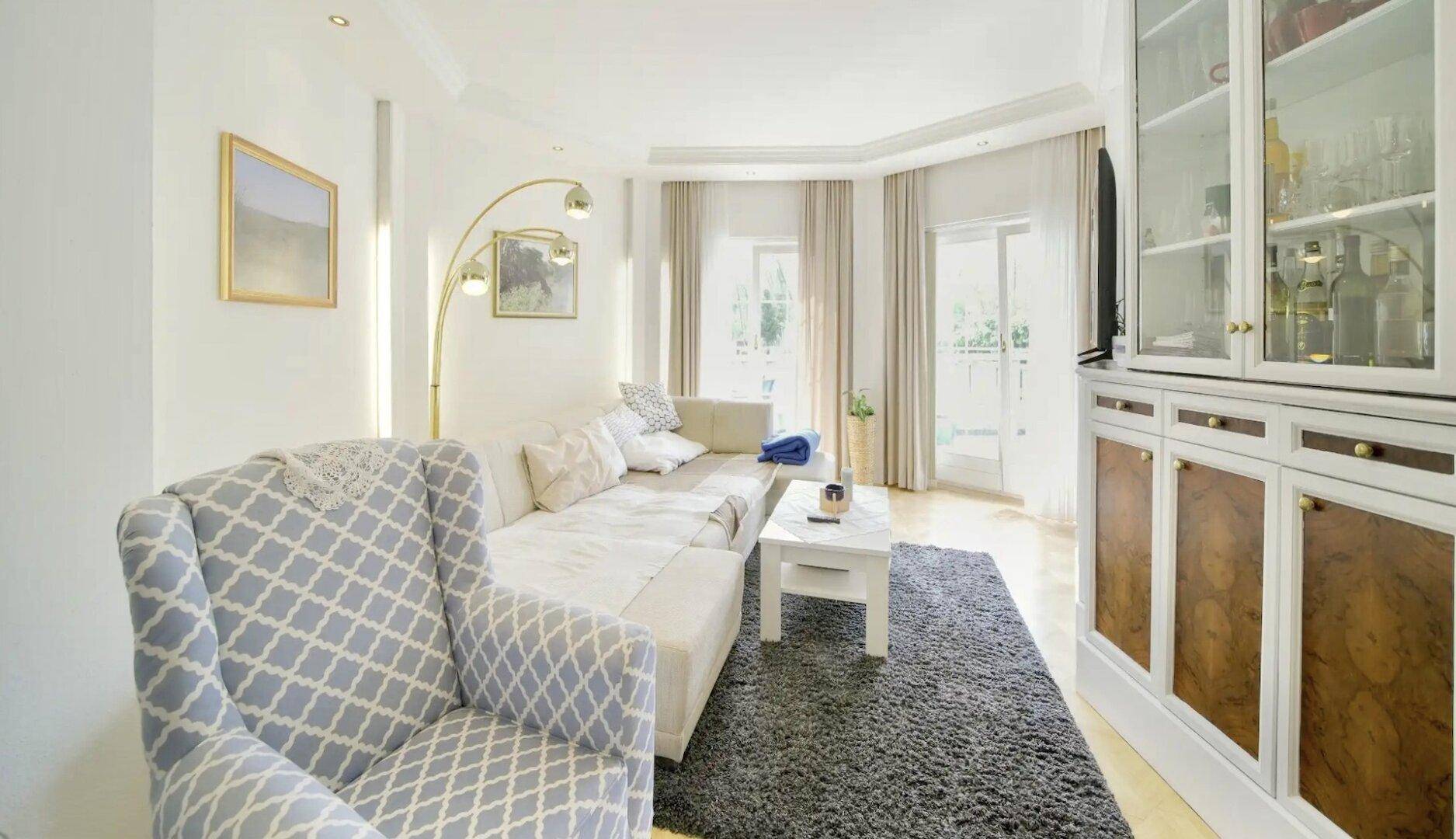 Gemütliches Wohnzimmer mit bequemer Couch, Teppich und großen Fenstern für viel Tageslicht.