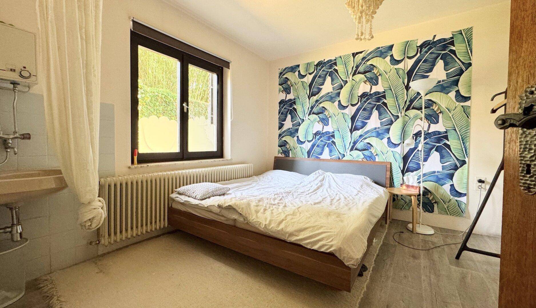 Schlafzimmer mit auffälliger Tapete, Holzbett und Fenster mit Blick ins Grüne.