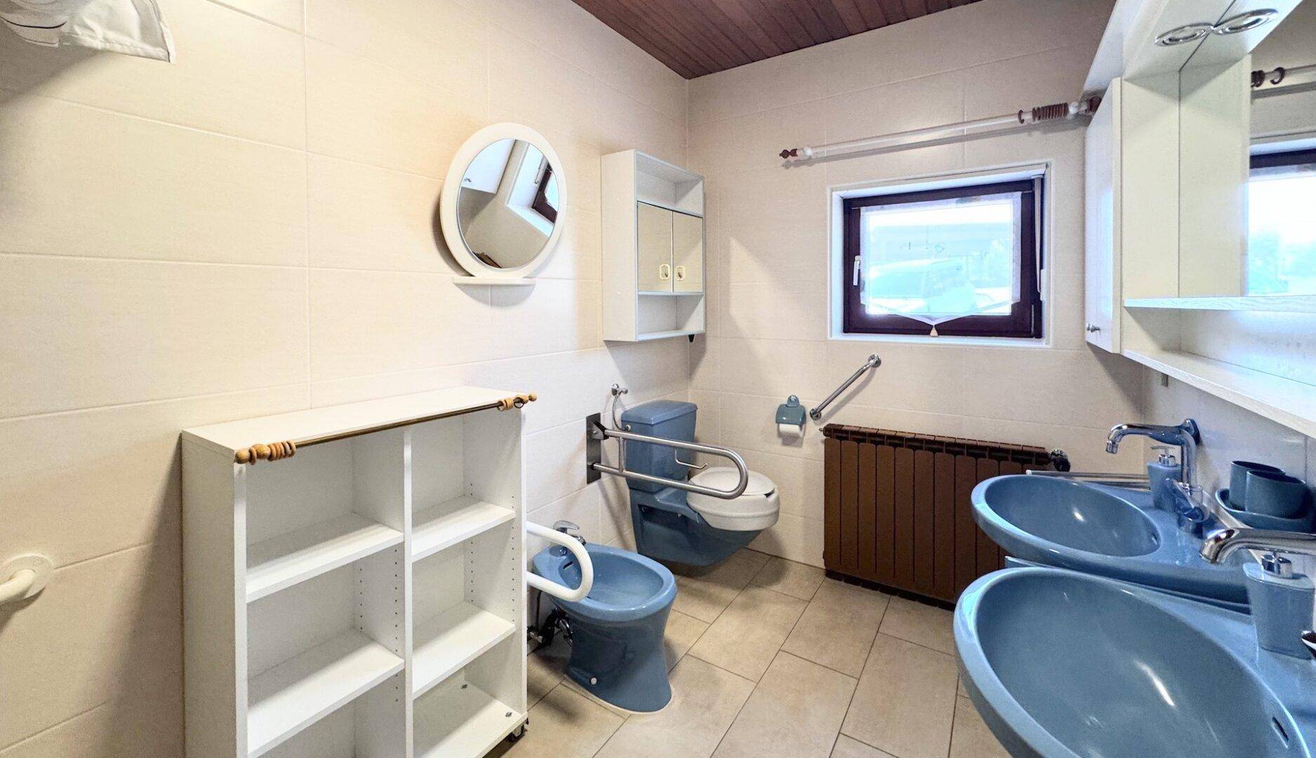 Badezimmer mit blauen Sanitäranlagen, Fliesenboden und Haltegriffen für Barrierefreiheit.