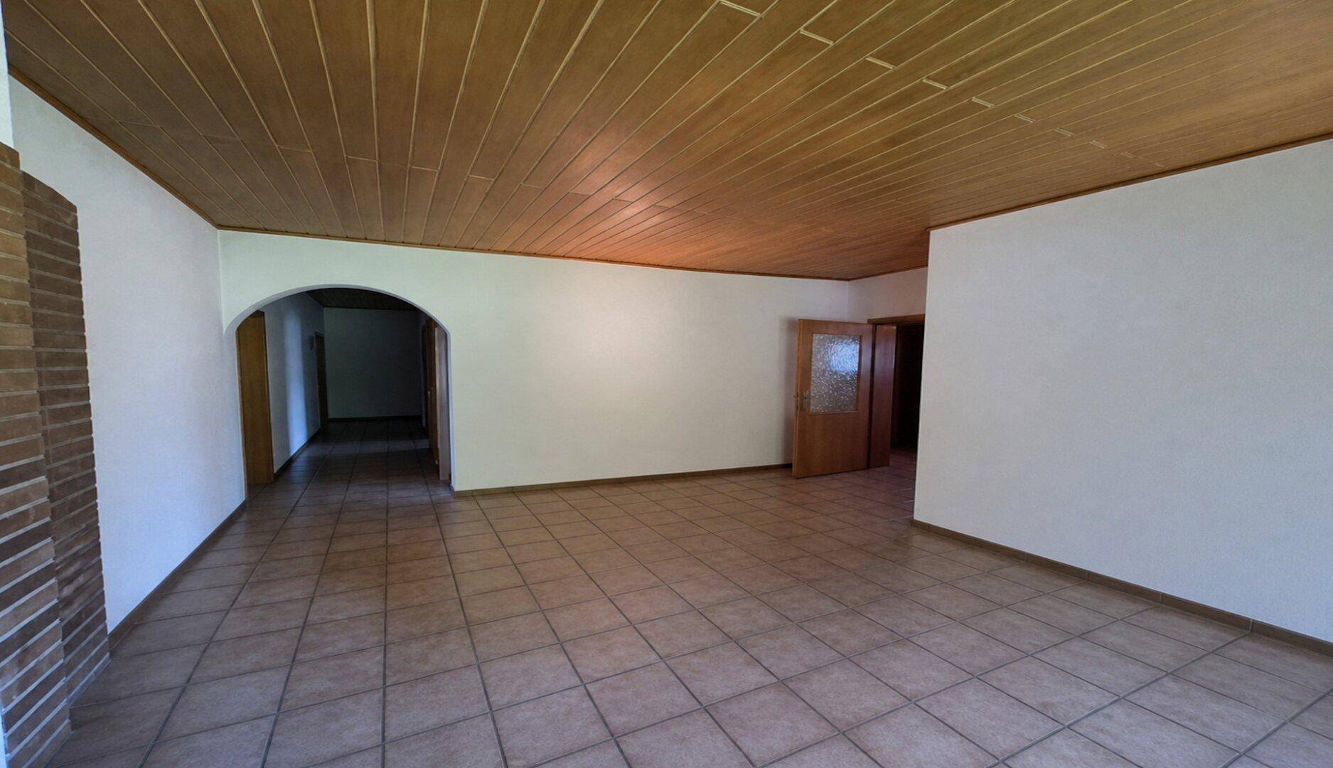 Geräumiger Wohnbereich mit Fliesenboden, Holzdecke und Zugang zu weiteren Räumen durch einen Bogen.