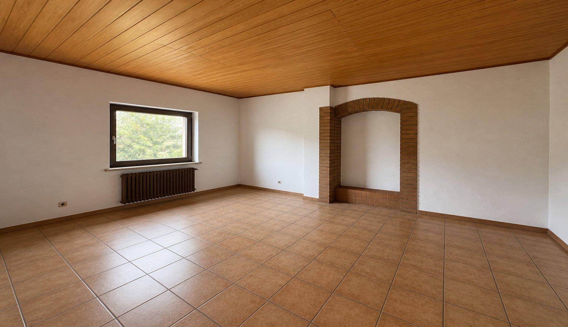 Leerer Raum mit Fliesenboden, Holzdecke und einem gemauerten Kaminbereich unter dem Fenster.