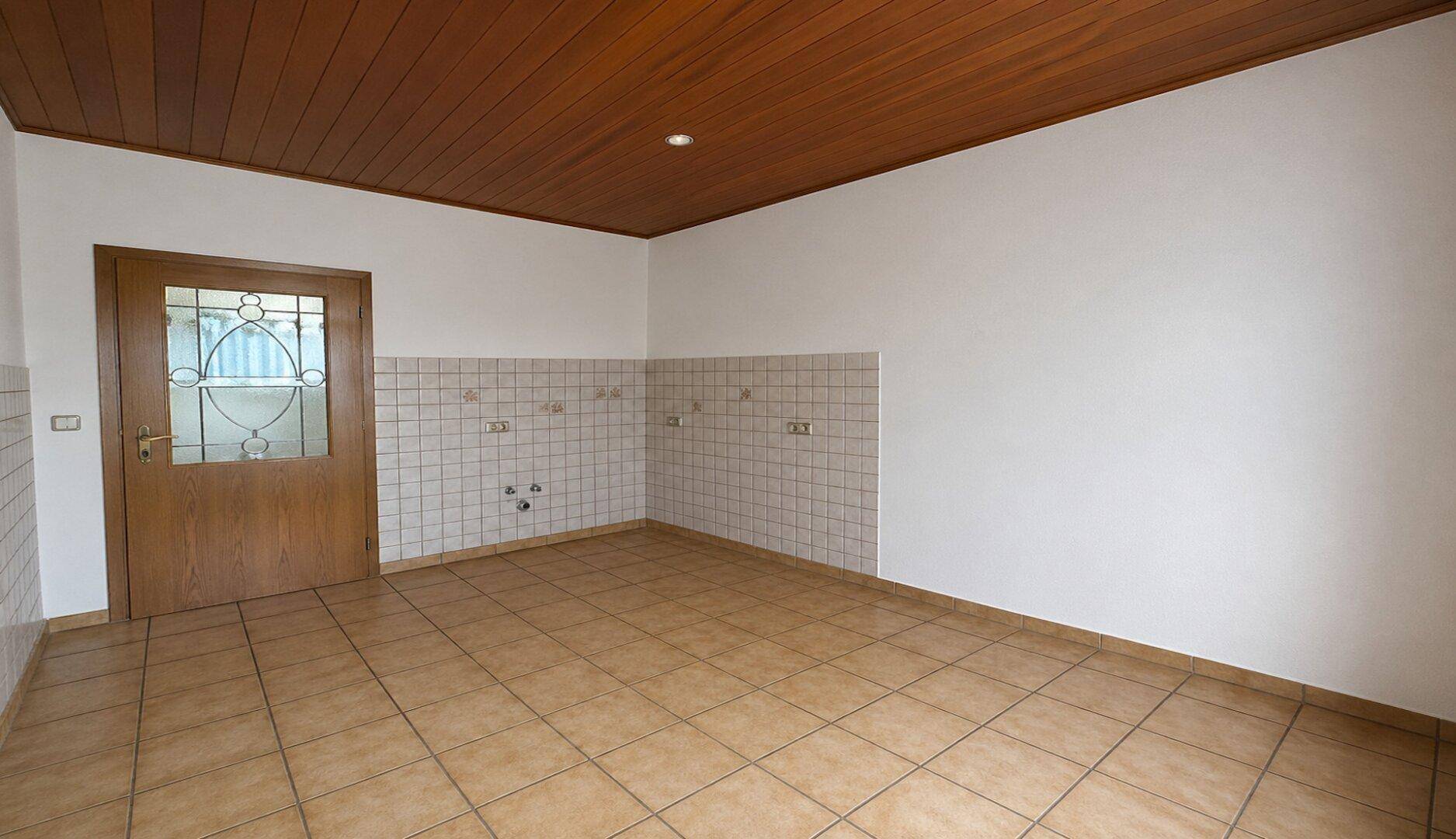 Leere Küche mit gefliesten Wänden, Holzdecke und Anschlussmöglichkeiten für Küchengeräte.