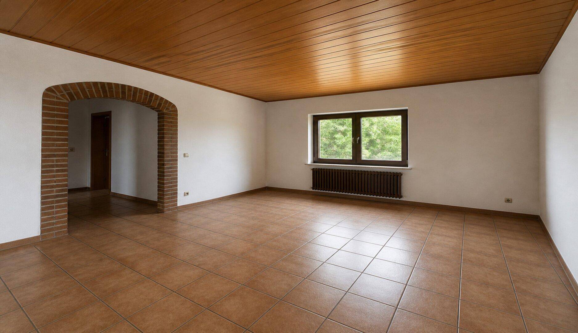 Heller, leerer Raum mit Fliesenboden, Holzdecke und einem gemauerten Bogen zu einem weiteren Bereich.