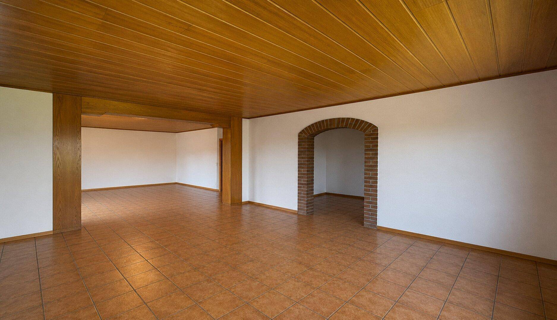 Großer offener Wohnbereich mit Fliesenboden, Holzdecke und sichtbaren Holzbalken.