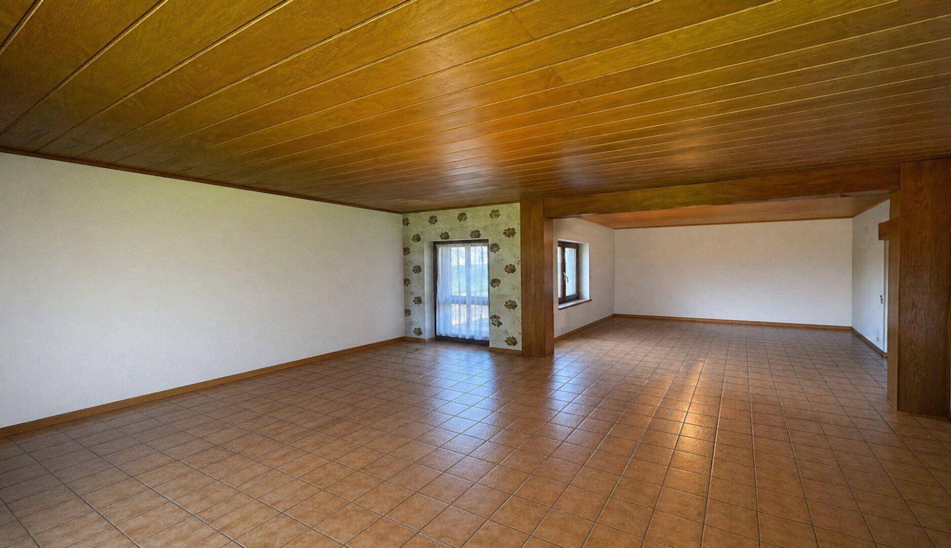 Geräumiger Wohnbereich mit Fliesenboden, Holzdecke und Zugang zu einem Außenbereich.