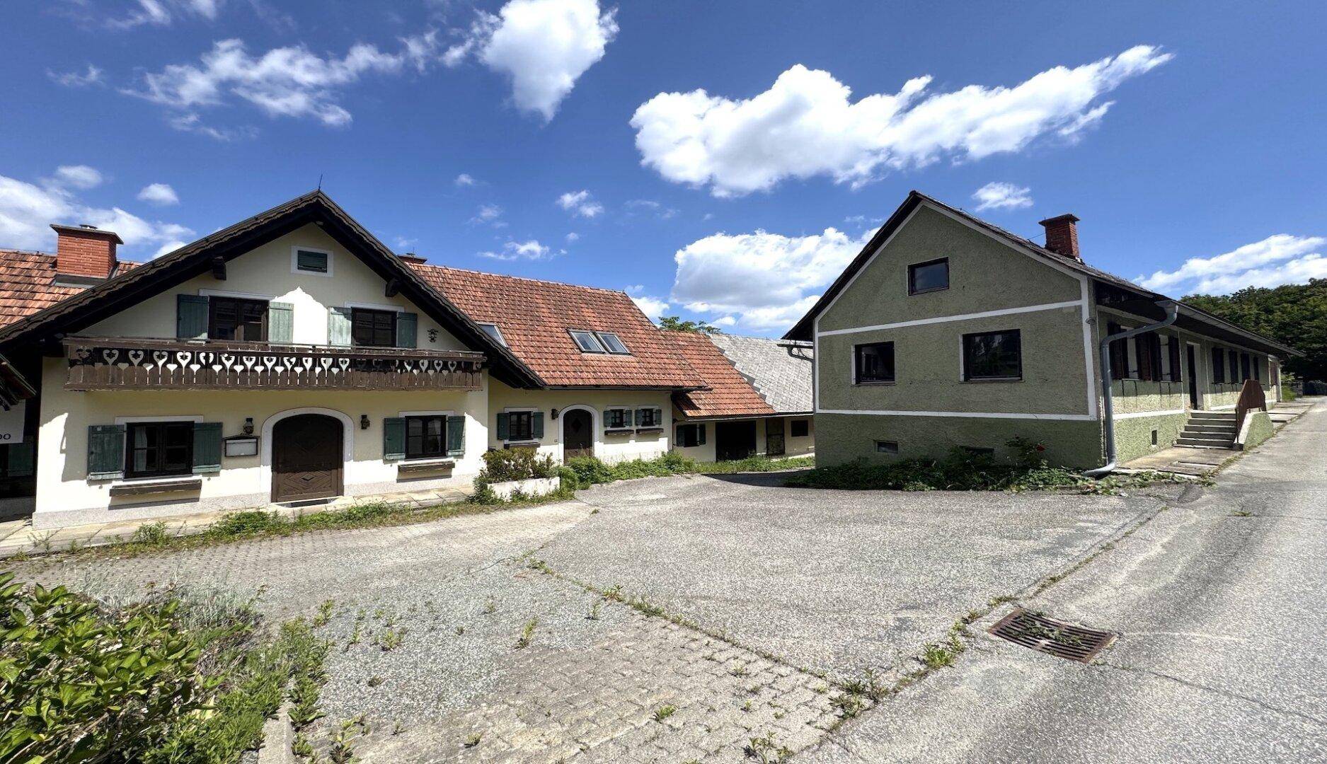 Zwei Gebäude mit unterschiedlicher Architektur und einem gepflasterten Vorplatz in ländlicher Umgebung.