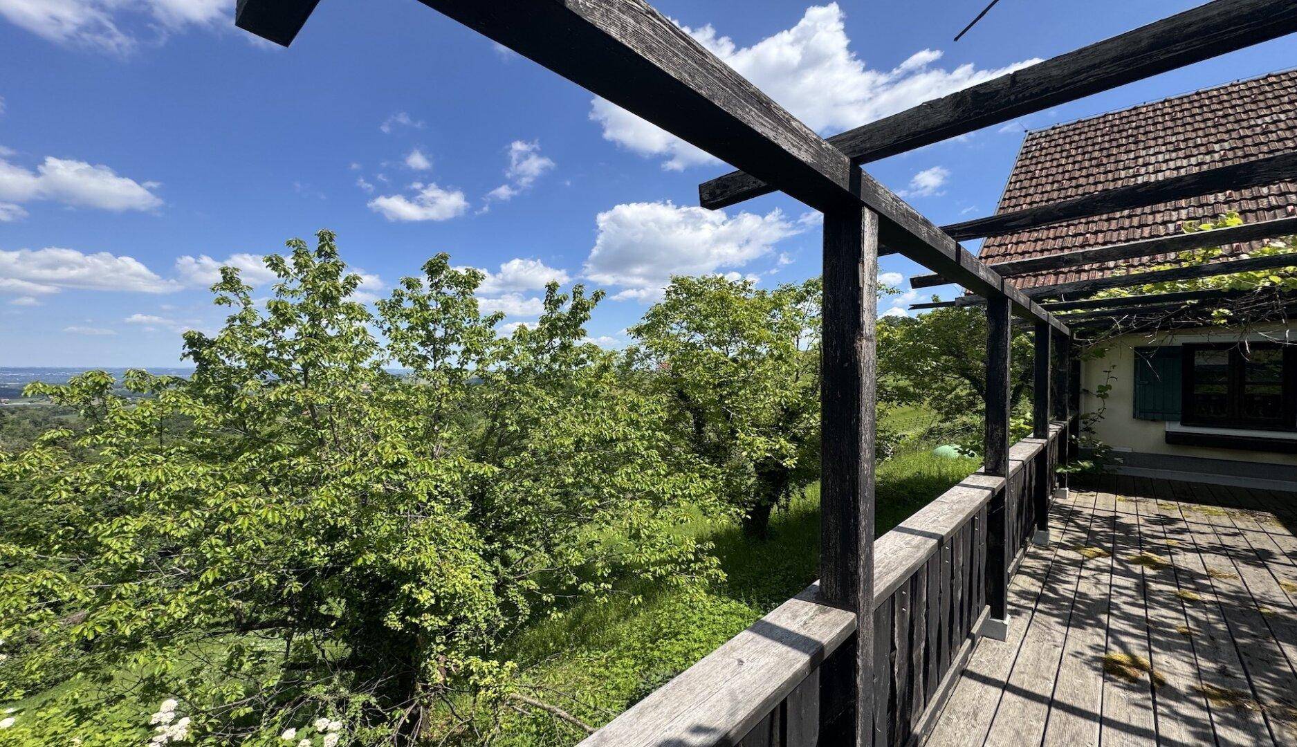 Weitblick von einer Holzterrasse mit Pergola über grüne Bäume und die umliegende Landschaft.