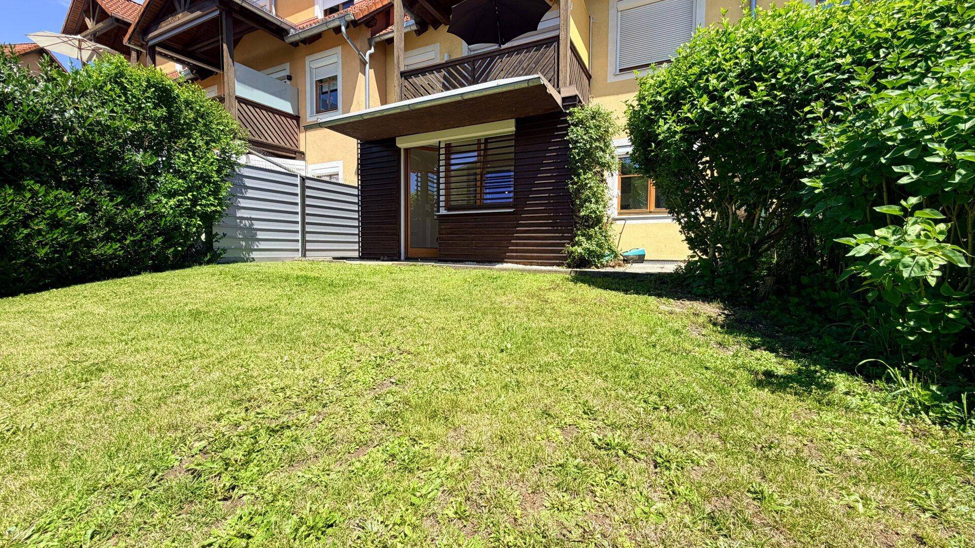 Sonniger Garten mit Rasenfläche und Zugang zu einer überdachten Terrasse im Erdgeschoss.