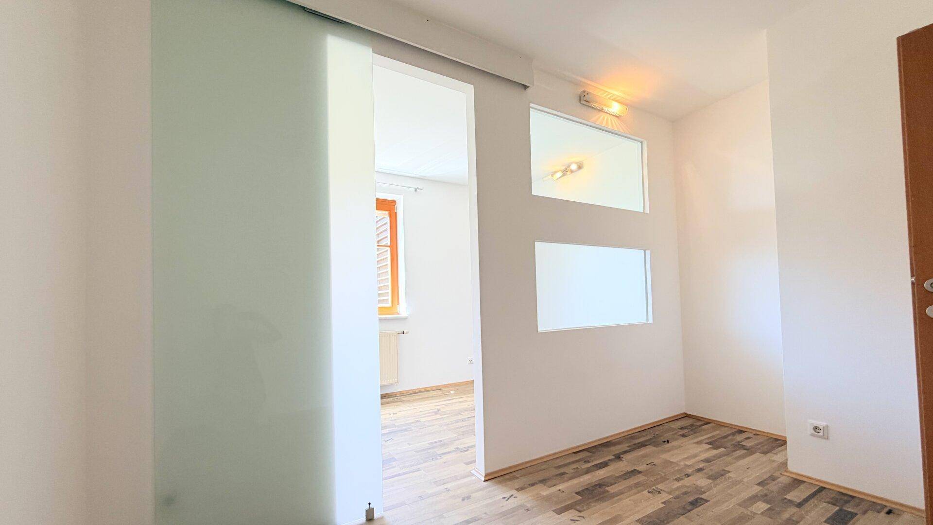 Raum mit Holzboden und einer modernen Glasschiebetür, die zu einem weiteren Zimmer führt.