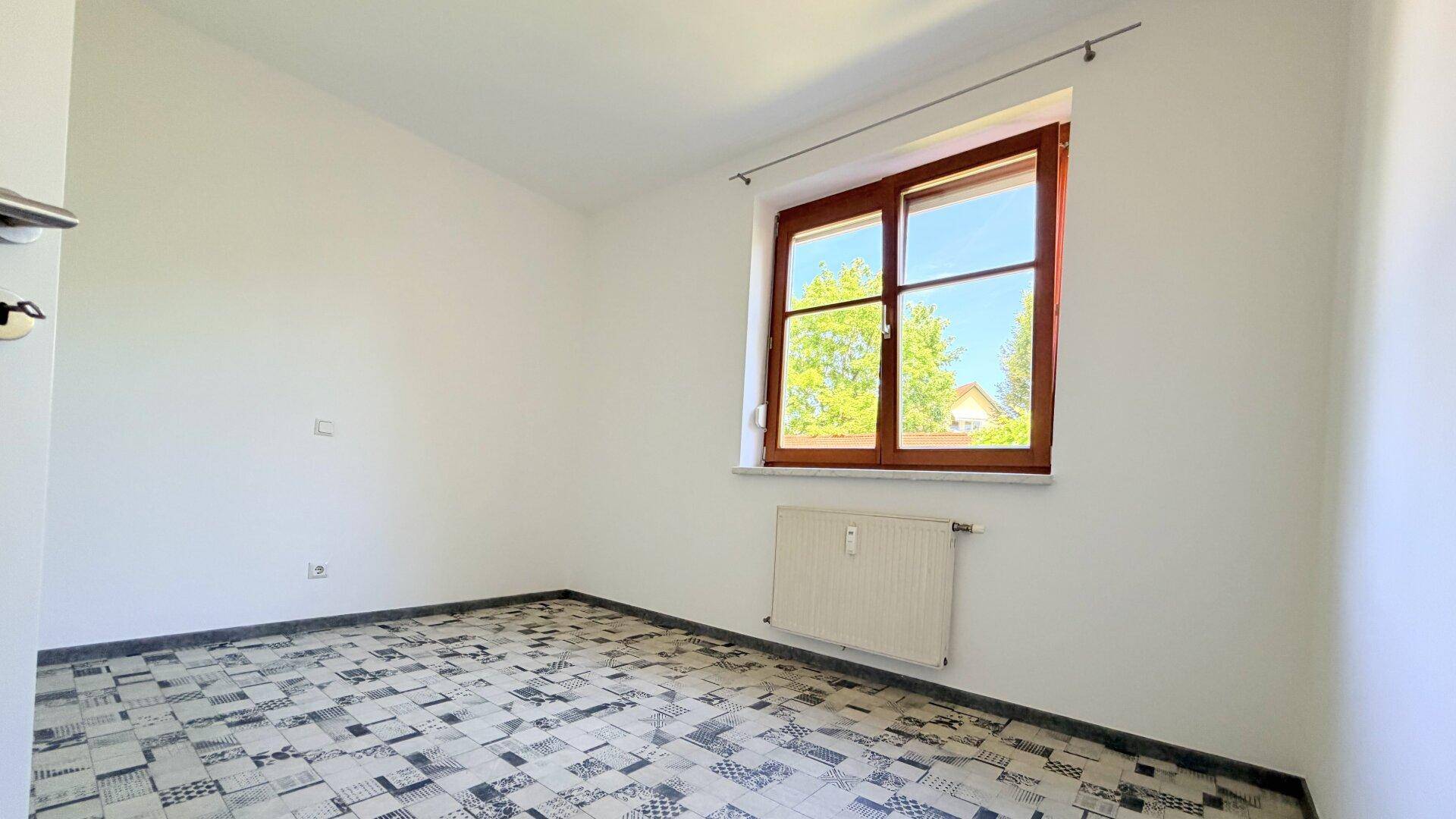 Helles Schlafzimmer mit gemustertem Bodenbelag und einem Fenster mit Blick ins Grüne.