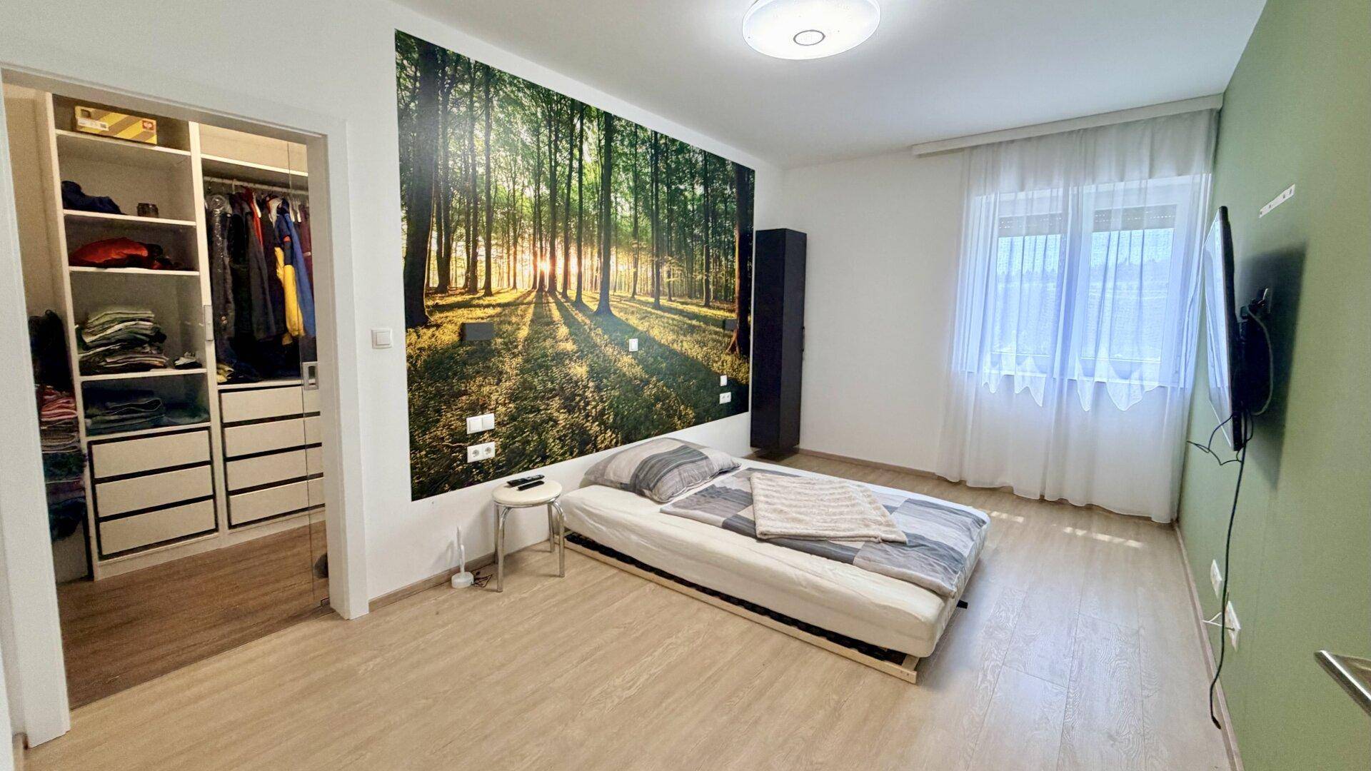 Schlafzimmer mit Bett, Wald-Wandbild und offenem begehbarem Kleiderschrank.