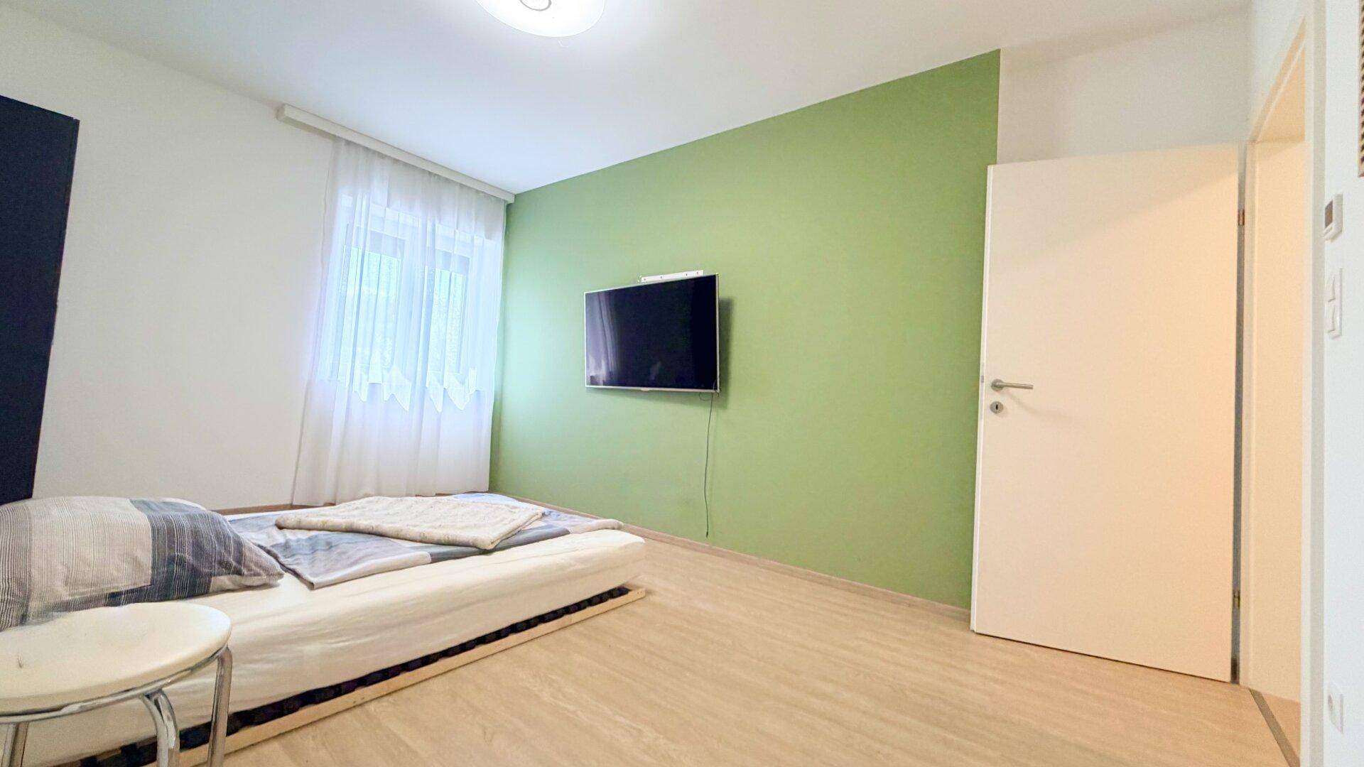 Schlafzimmer mit grün gestrichener Wand, Fernseher und hellem Laminatboden.