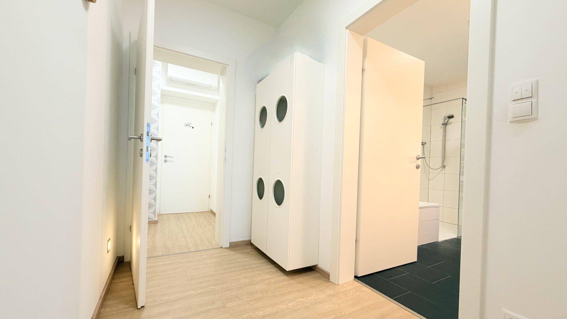 Heller Flur mit weißem Schrank und Blick in ein Badezimmer mit Dusche.