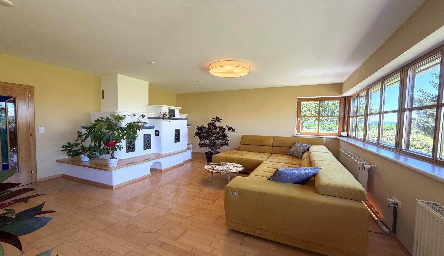 Helles Wohnzimmer mit großem Kachelofen, gelbem Ecksofa und weitem Ausblick.