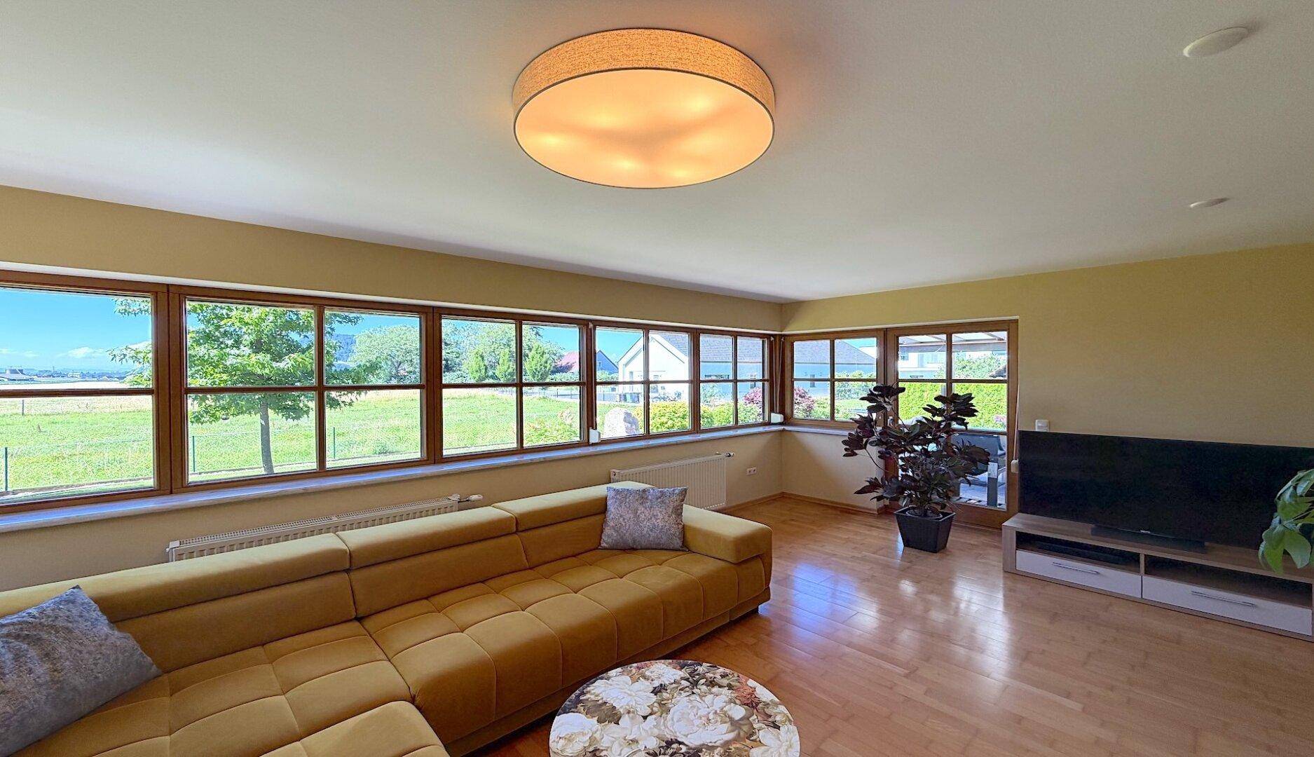 Geräumiges Wohnzimmer mit großzügiger Fensterfront, Holzboden und Blick ins Grüne.