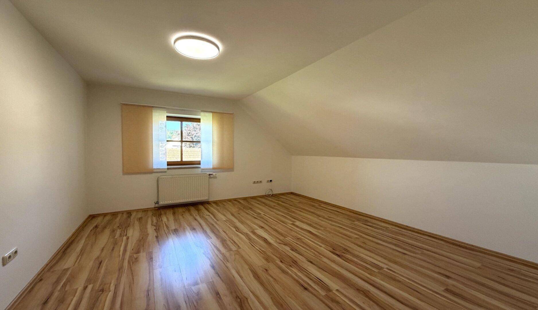 Helles Zimmer mit Holzboden, Dachschräge und Fenster mit Blick nach draußen.
