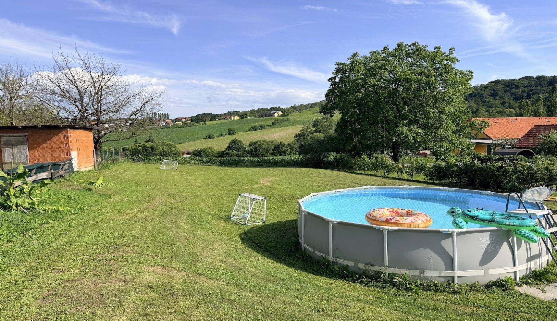 Sonniger Garten mit Aufstellpool, kleinen Fußballtoren und Blick auf die grüne Umgebung.