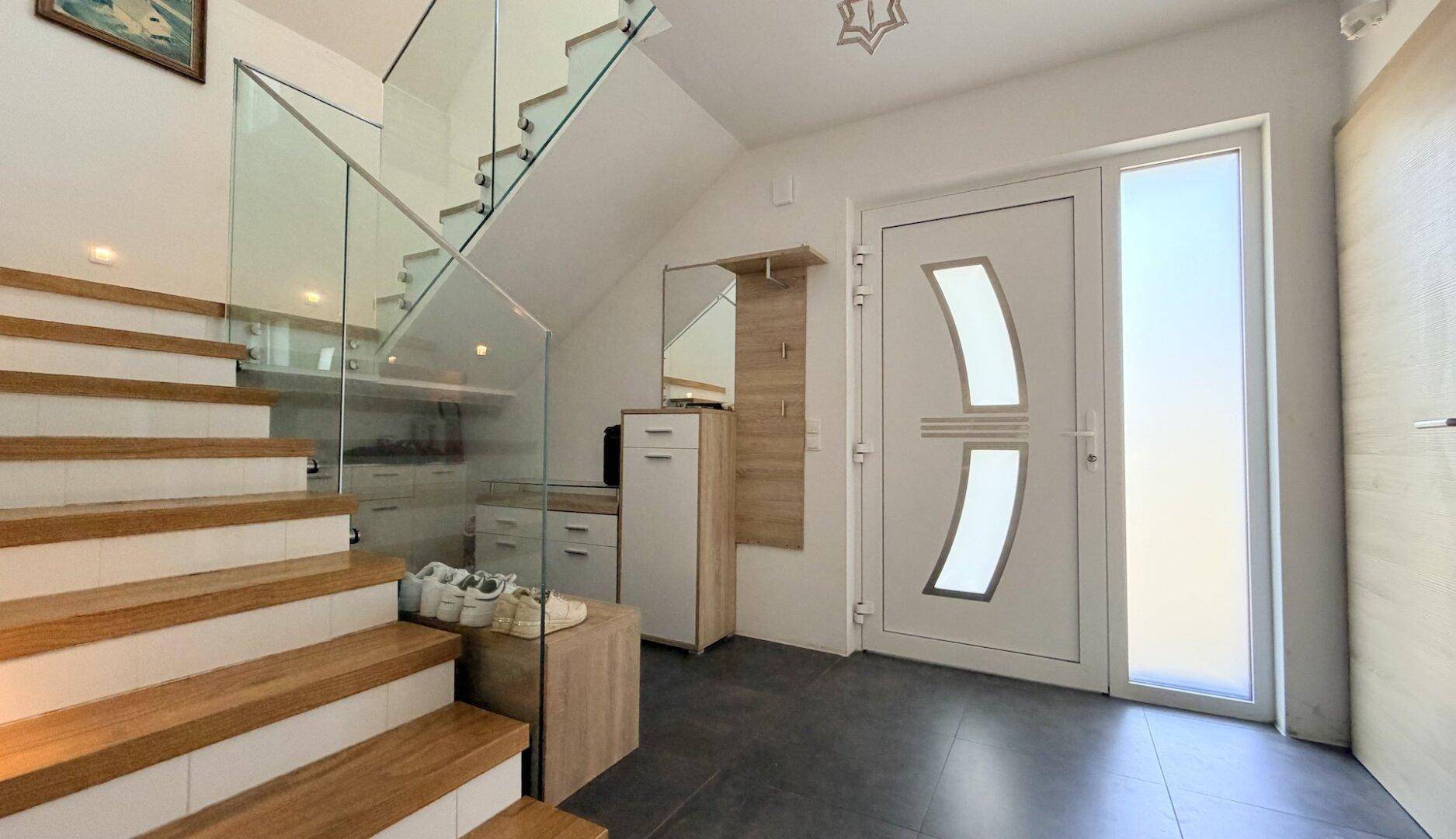 Heller Eingangsbereich mit moderner Holztreppe, Glasgeländer und dunklen Fliesen.