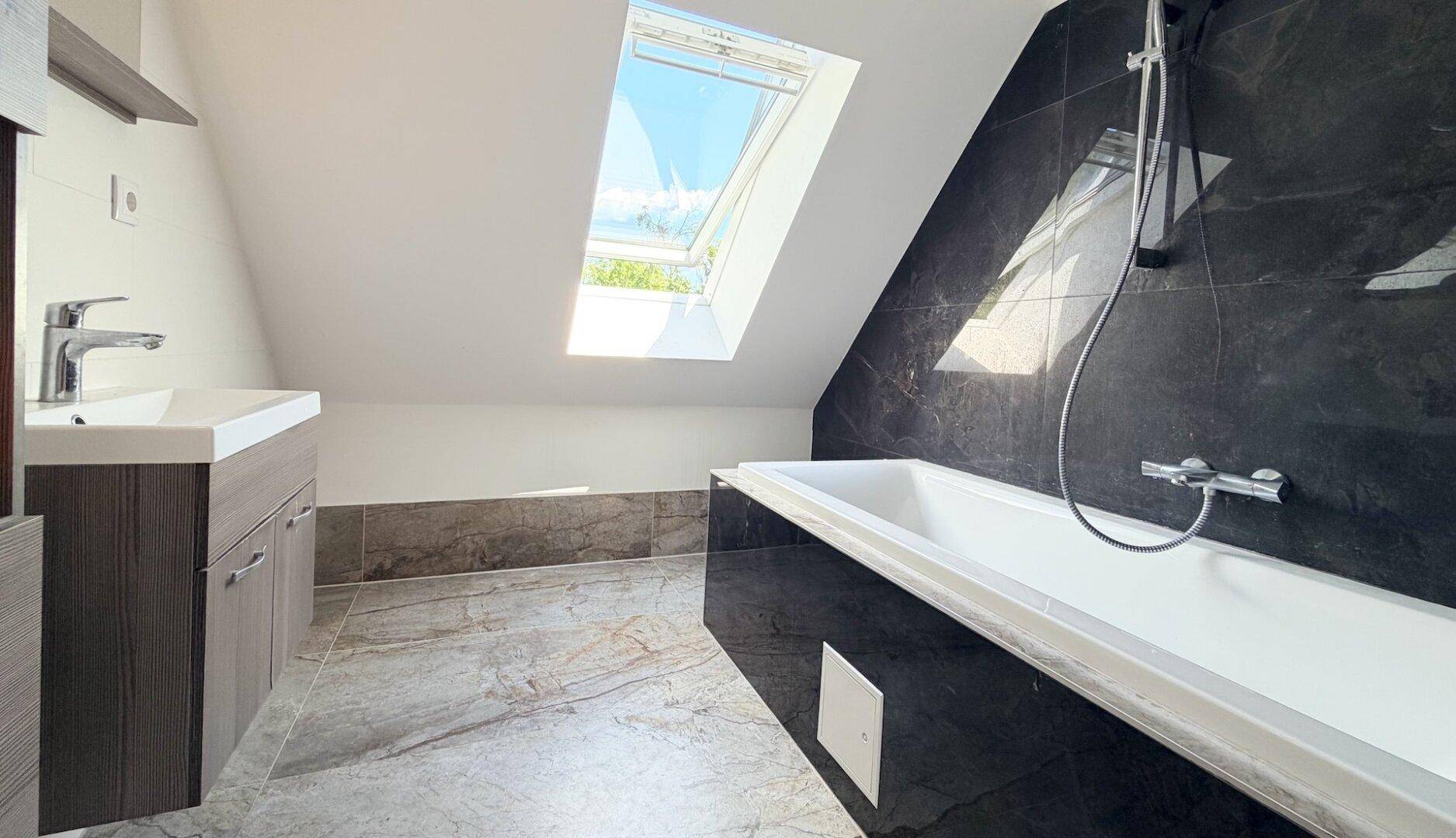 Modernes Badezimmer im Dachgeschoss mit Badewanne, dunklen Fliesen und Dachfenster.