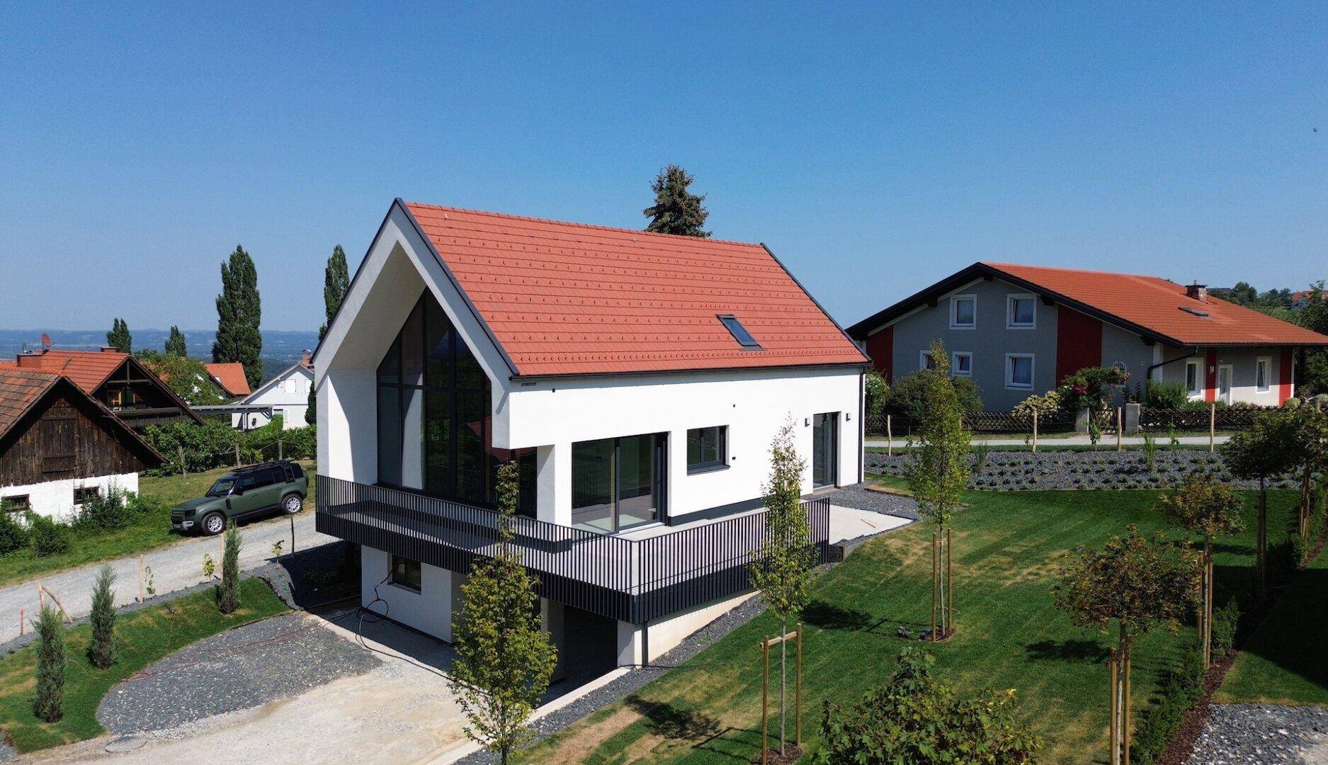 Modernes Einfamilienhaus mit rotem Ziegeldach und Balkon, umgeben von grüner Landschaft und Seeblick.