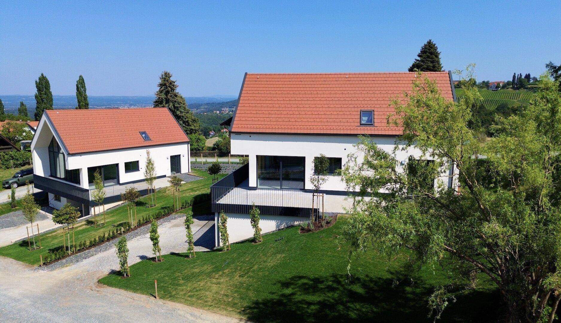 Zwei moderne Häuser mit ähnlicher Architektur, großen Glasfronten und gepflegten Gärten in ländlicher Lage.