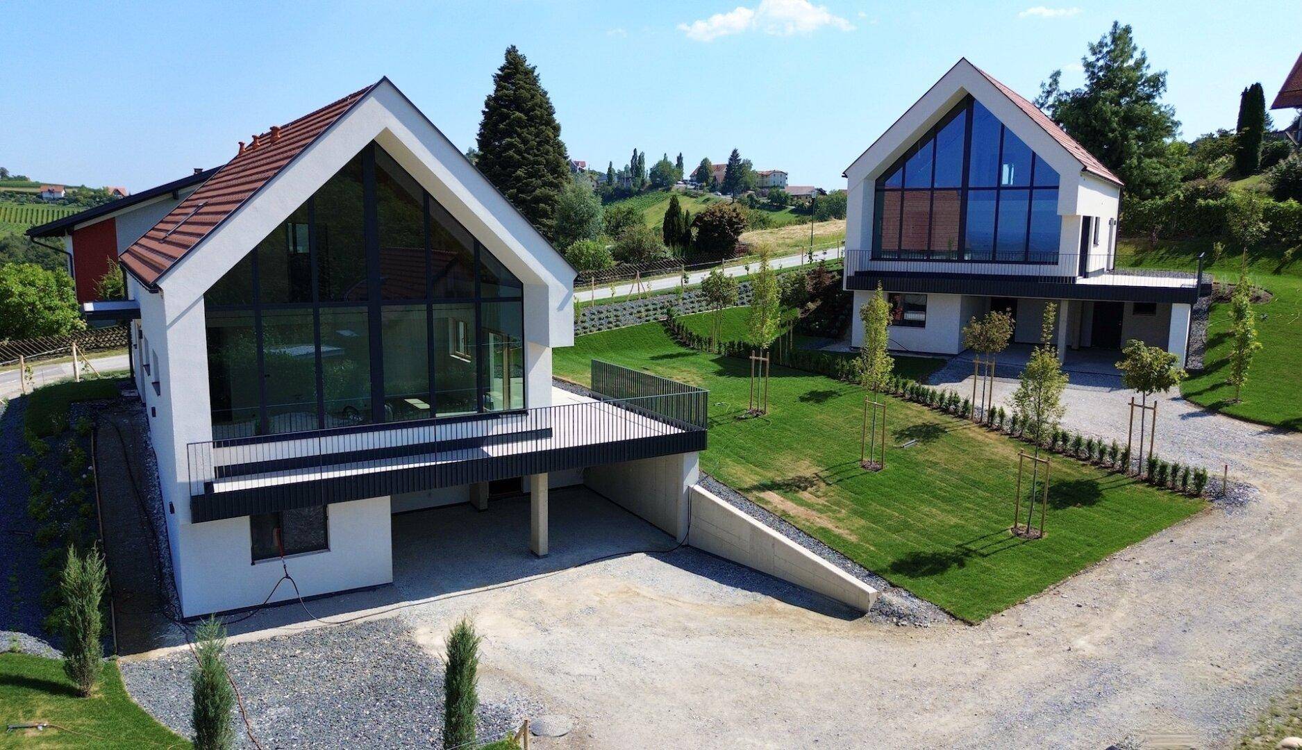 Zwei moderne Häuser mit großen Glasfronten und Balkonen, umgeben von gepflegten Gärten und Zufahrten.
