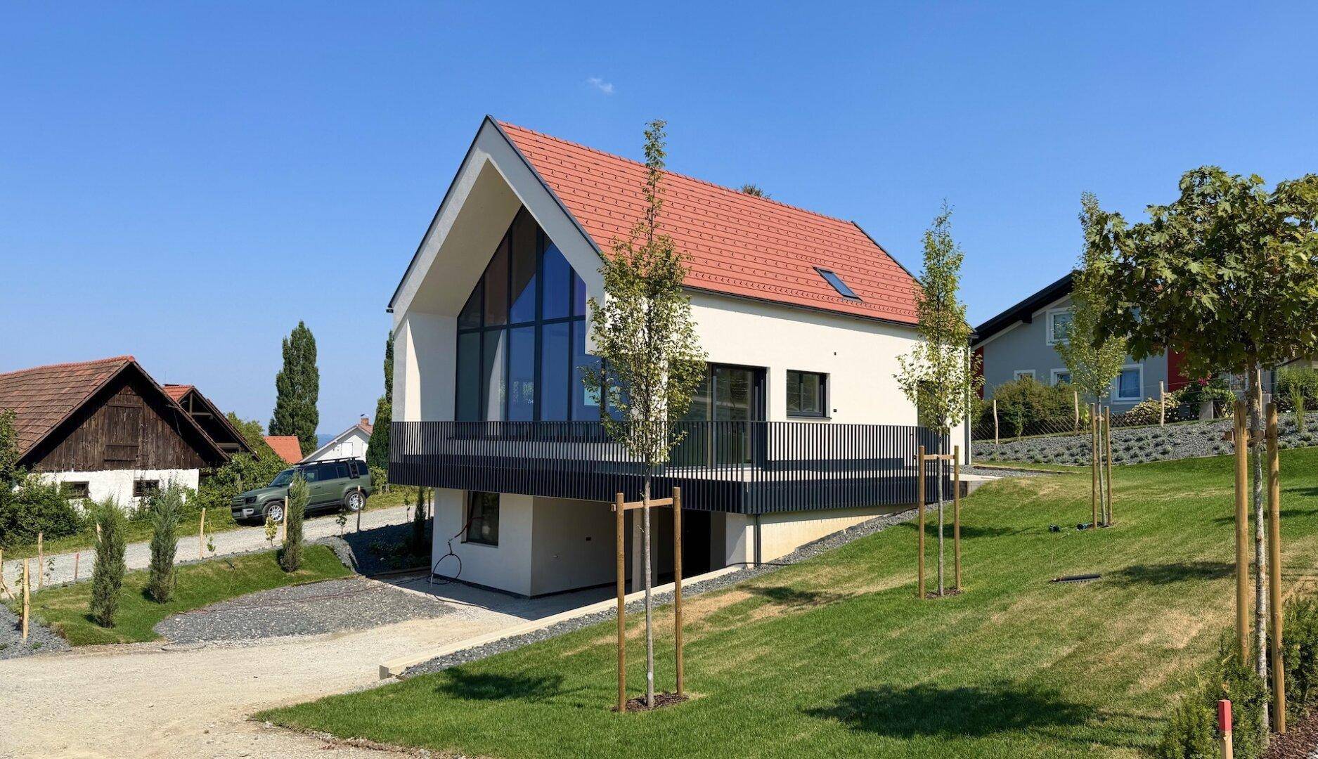 Modernes Haus mit Balkon und großer Glasfront, umgeben von jungem Grün und einer Schotterzufahrt.