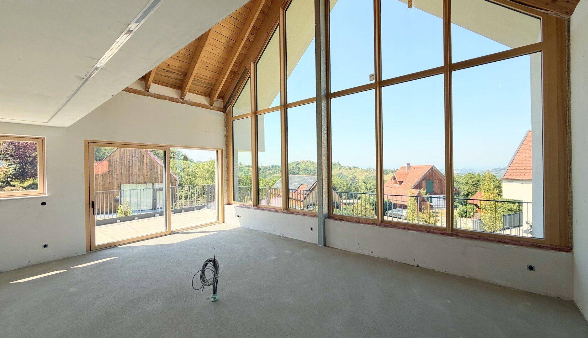 Lichtdurchfluteter Wohnbereich im Rohbau mit Holzbalkendecke, großer Glasfront und Zugang zu einem Außenbereich.