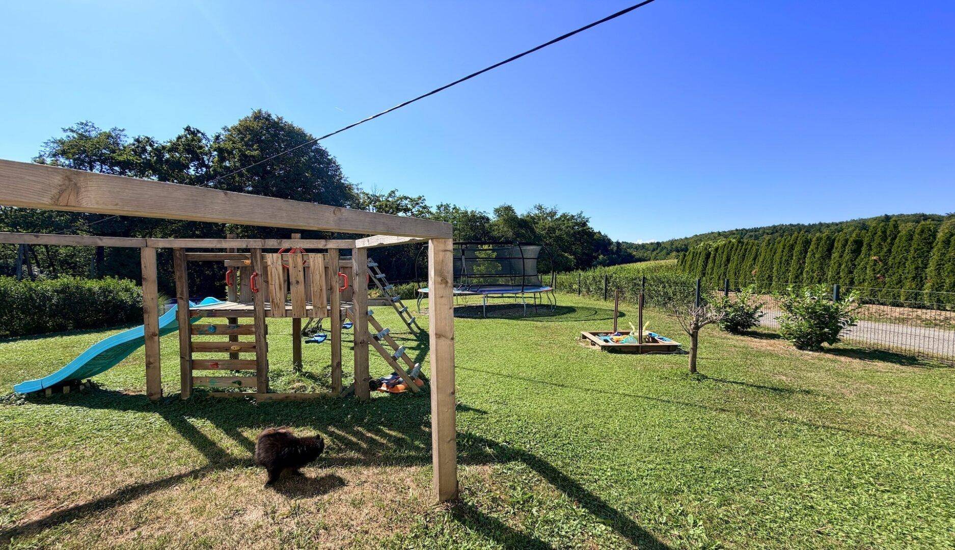 Sonniger Garten mit großem Holzspielplatz, Sandkasten und Trampolin für Kinder.