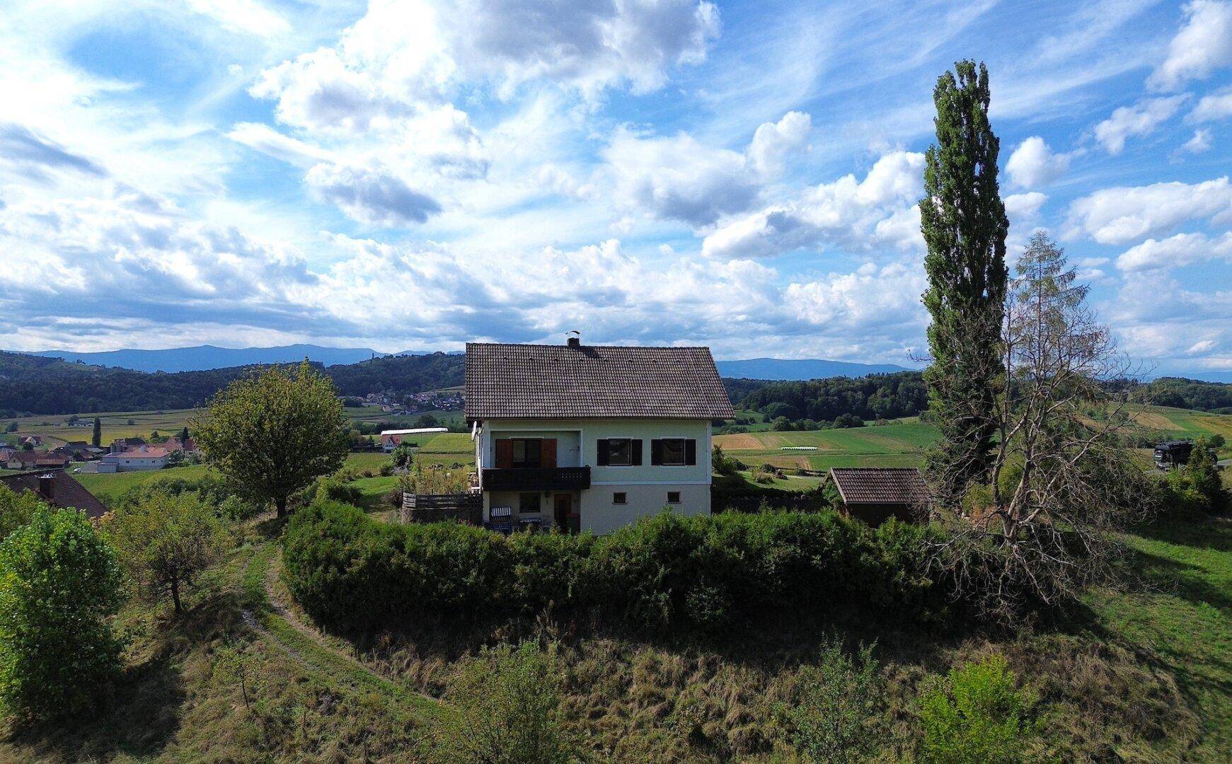 Freistehendes Haus mit Balkon und grüner Hecke, umgeben von weitläufiger Naturlandschaft.