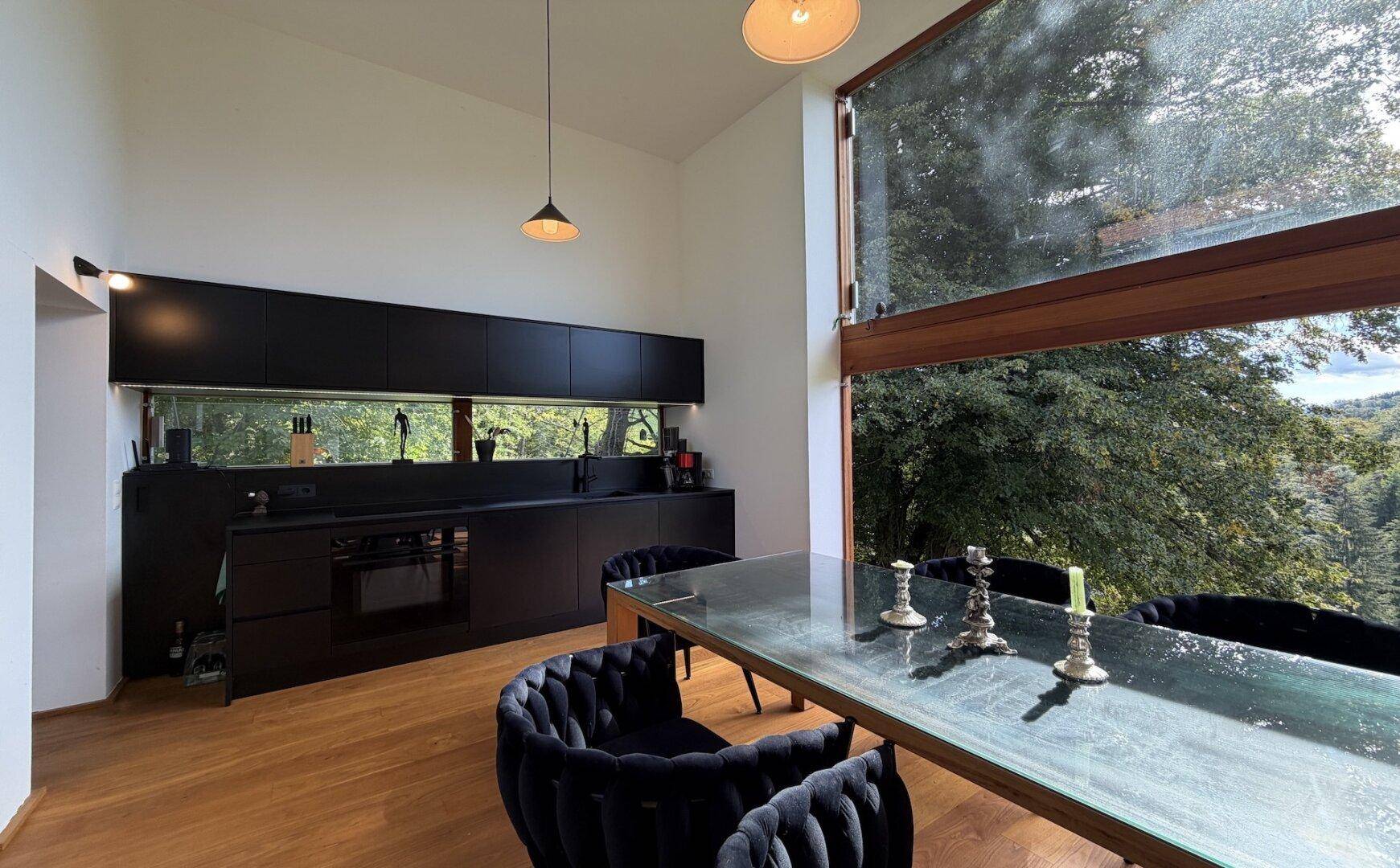 Elegante schwarze Küche mit Esstisch und Stühlen, umgeben von großen Fenstern mit Waldblick.