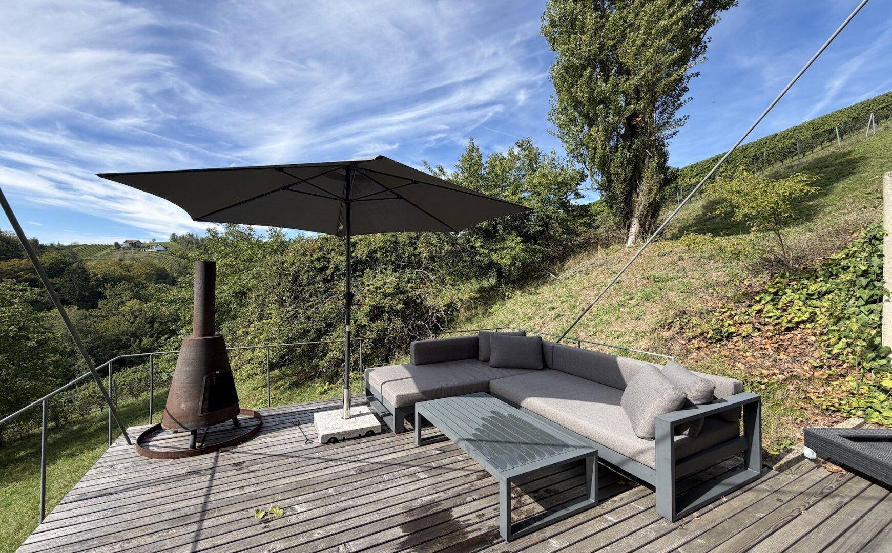 Große Holzterrasse mit bequemen Outdoor-Möbeln, Sonnenschirm und Feuerstelle mit Waldblick.