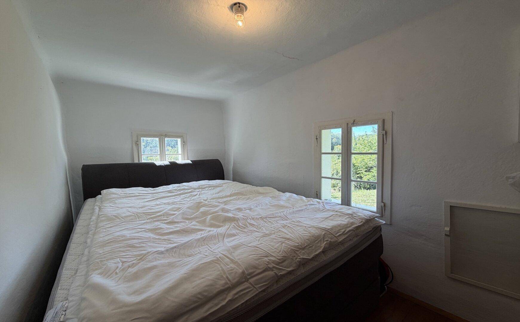 Helles Schlafzimmer mit Doppelbett, Holzboden und Fenstern, die viel Tageslicht hereinlassen.