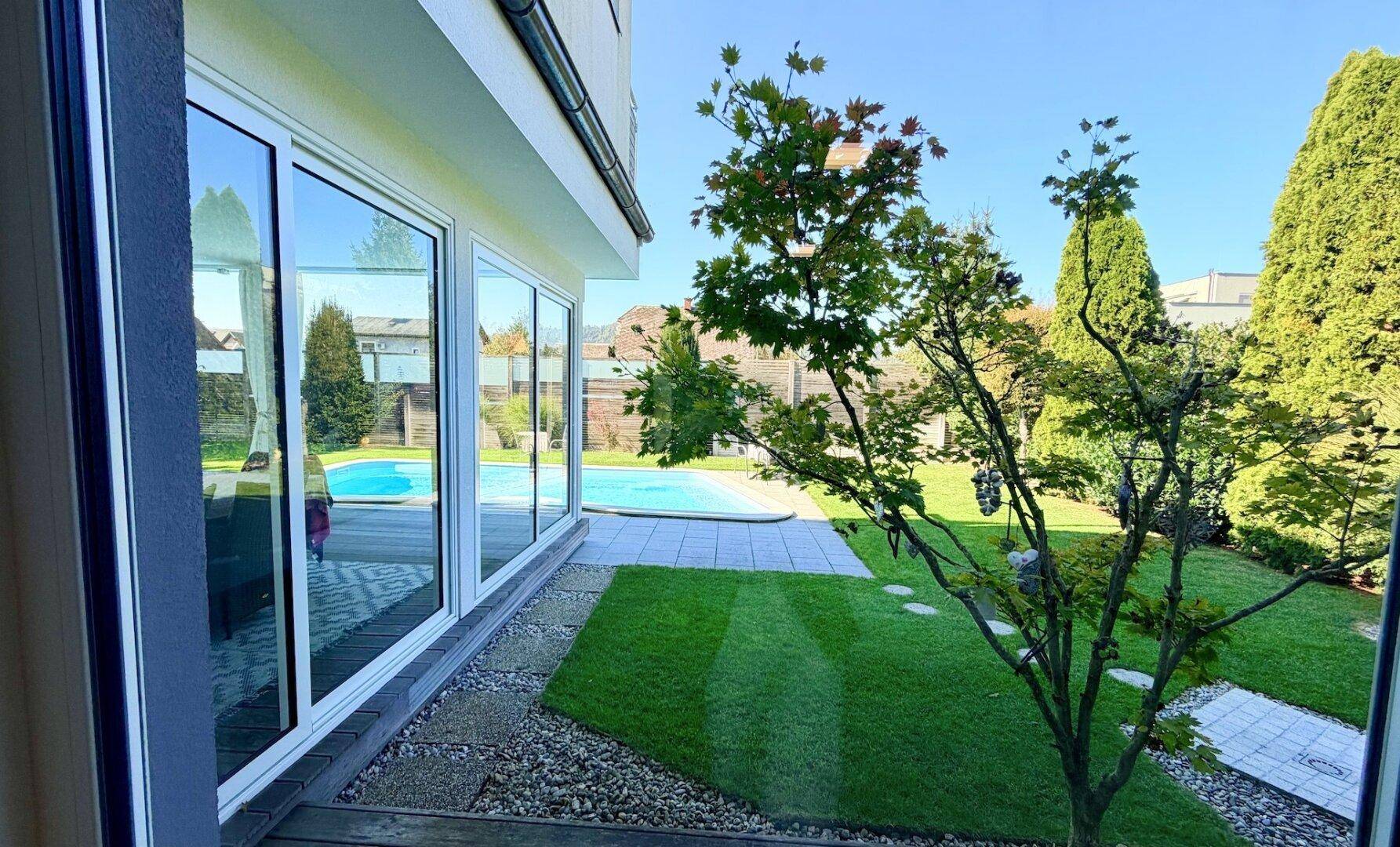 Heller Innenraum mit großen Fensterfronten und direktem Blick auf den Pool und Garten.