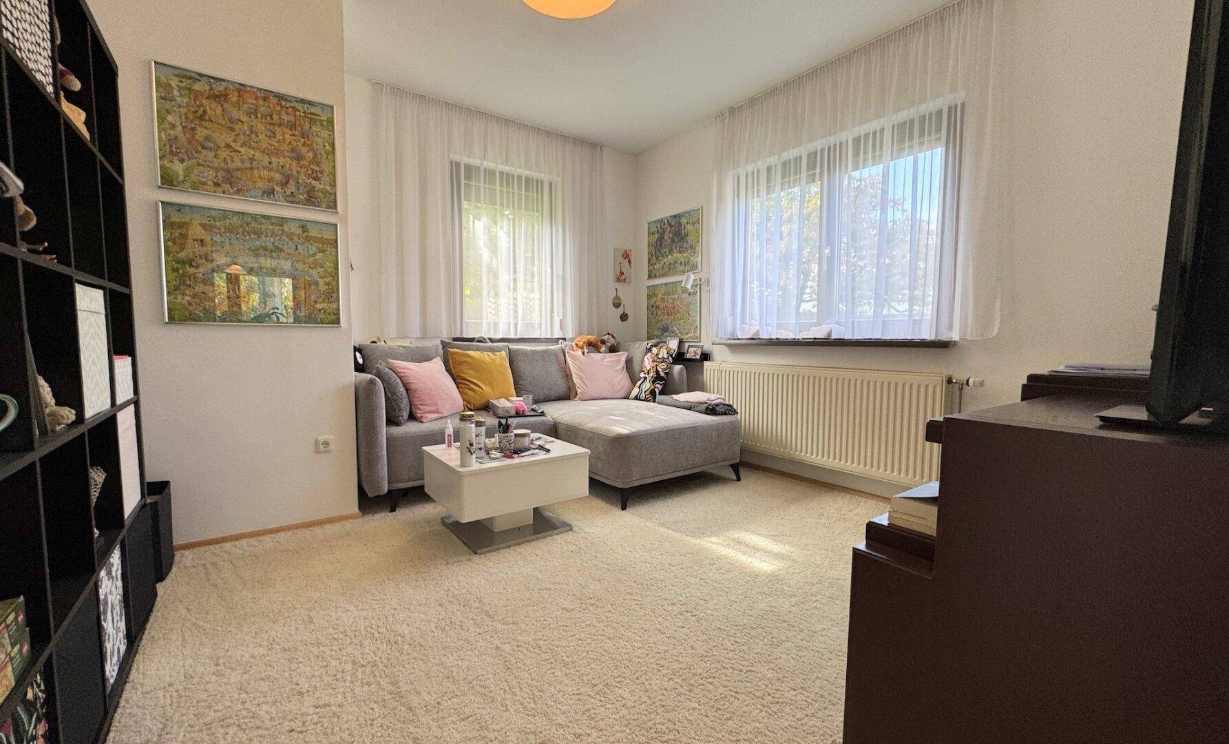 Helles Wohnzimmer mit gemütlichem Ecksofa, Teppichboden und großen Fenstern für viel Tageslicht.