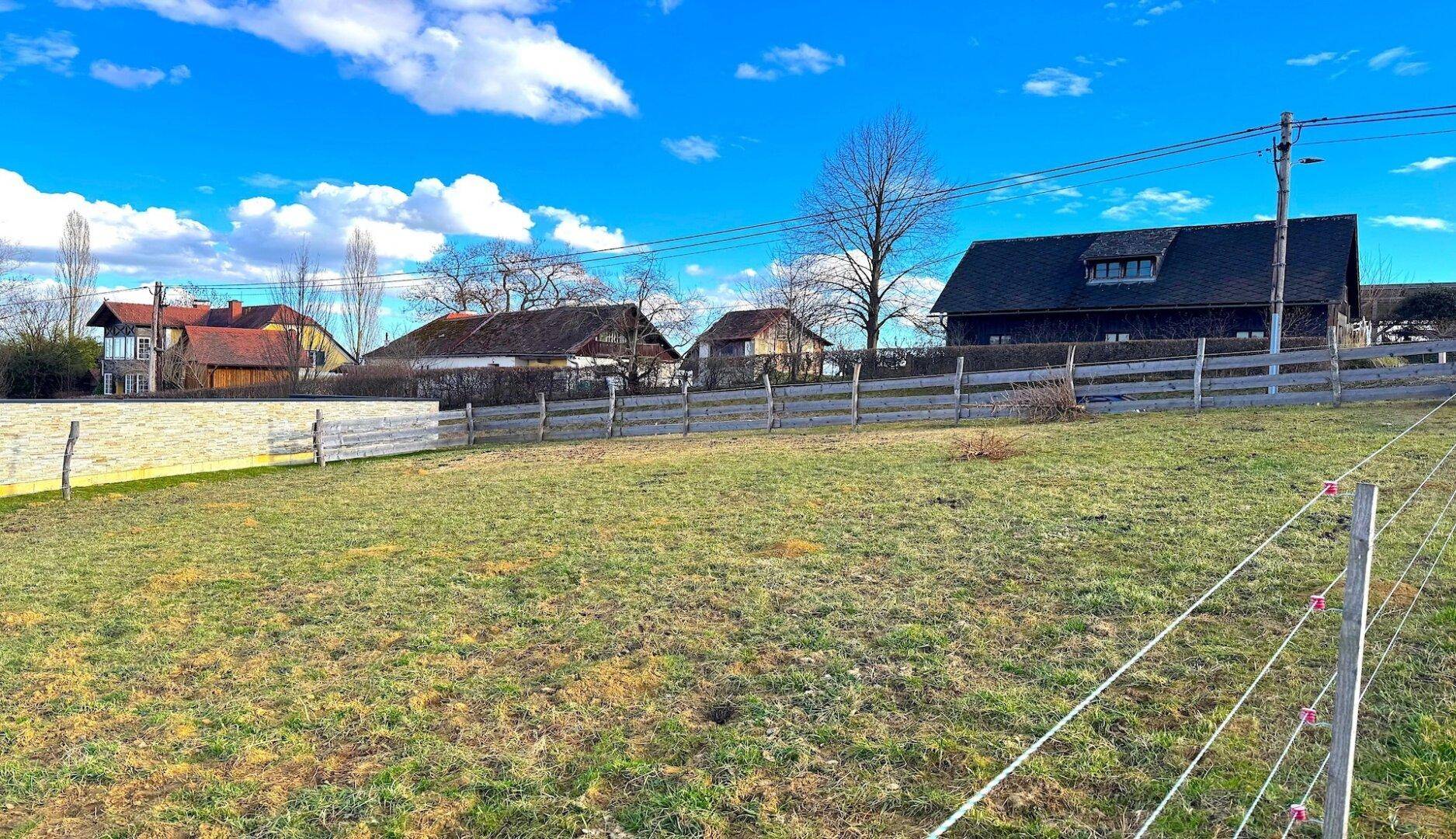 Offenes Feld mit einem Holzzaun und traditionellen Häusern im Hintergrund, umgeben von ländlicher Natur.
