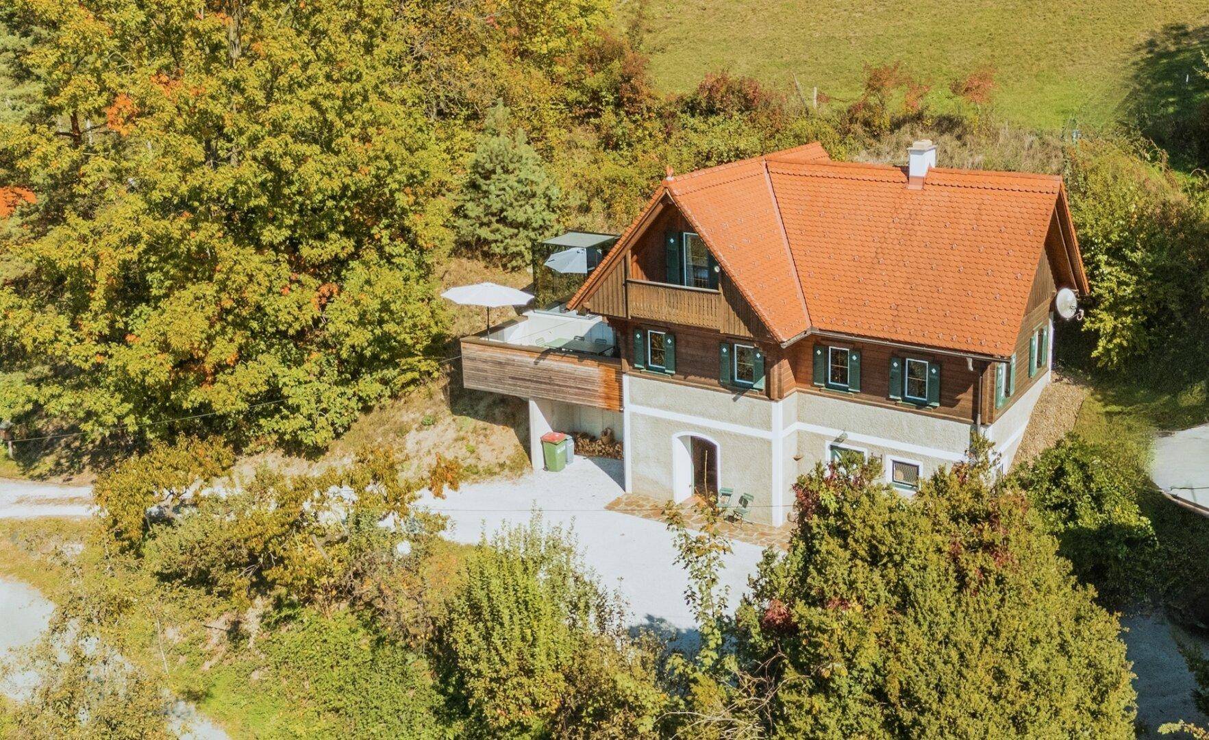 Landhaus in herbstlicher Landschaft mit umliegenden Bäumen und einer Terrasse mit Sonnenschirm.