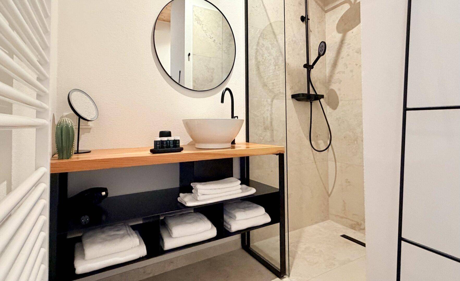 Stilvolles Badezimmer mit rundem Spiegel, schwarzer Armatur und begehbarer Dusche.