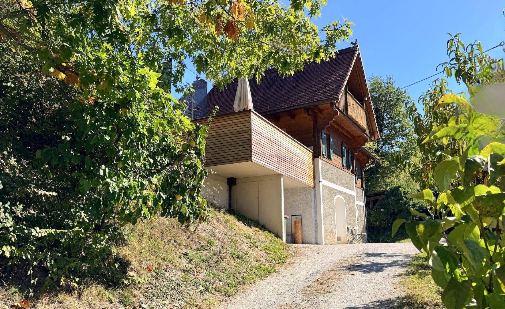 Landhaus mit moderner Holzterrasse, umgeben von üppiger Vegetation und einem Schotterweg.