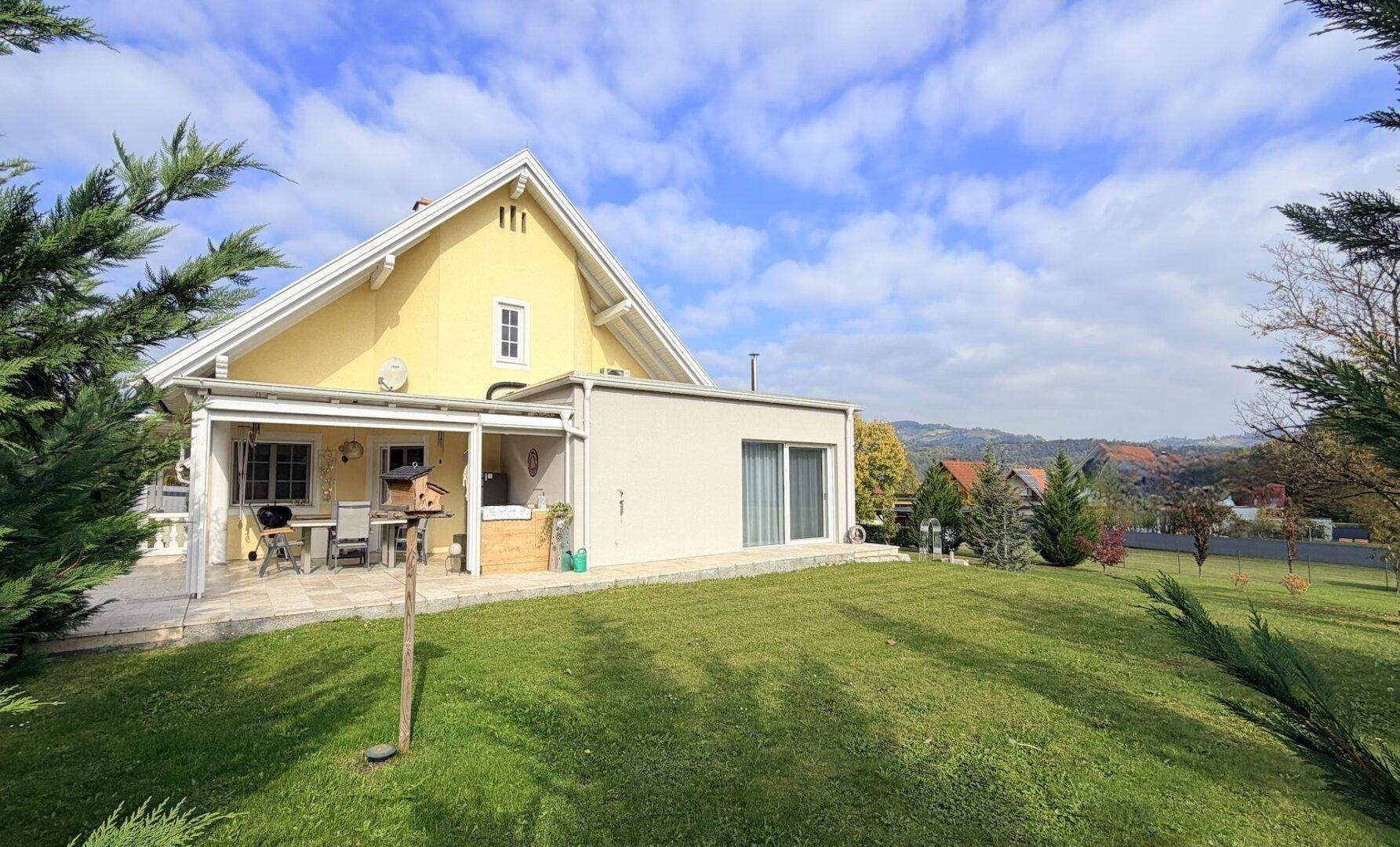 Rückseite des Hauses mit großer Rasenfläche und überdachter Terrasse unter blauem Himmel.