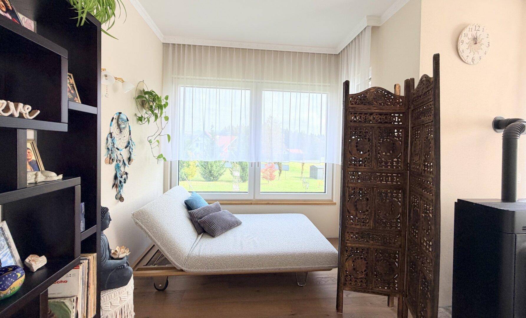 Heller Schlafbereich mit Tagesbett, Holzboden und Blick in den Garten durch das Fenster.